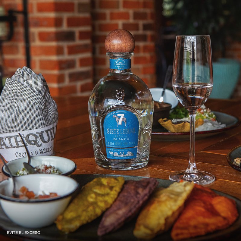 A bottle of "Siete Leguas Tequila Blanco" with a wooden cap, a wine glass with a clear liquid, and a plate of colorful tacos. Three small bowls of condiments and a brick wall background are also visible. The napkin holder reads "TEQUILA COCINA DE MEXICO."