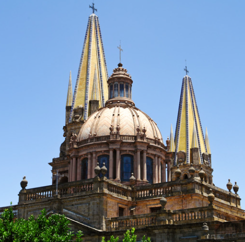 The Guadalajara Cathedral in Mexico with its central dome and four pointed spires, adorned with yellow and blue tiles against a clear blue sky.